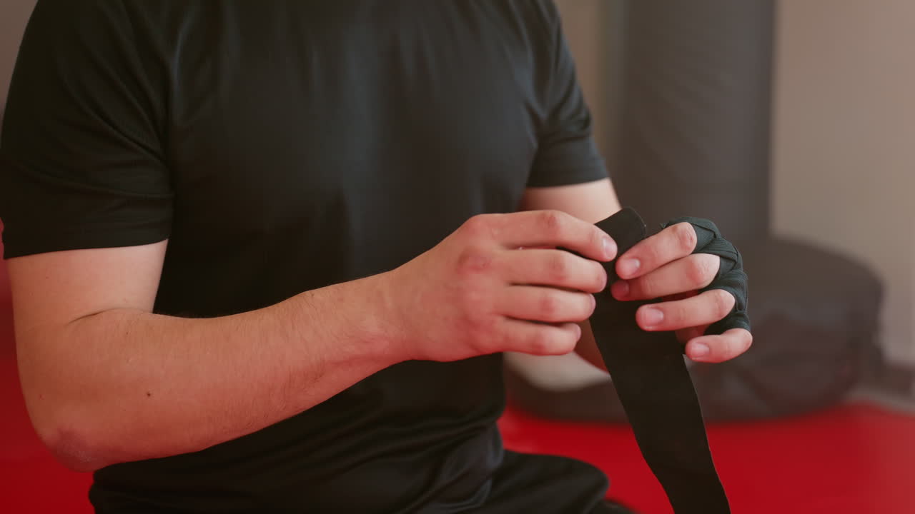 Athlete in black shirt sitting on red mat inside gym, preparing hand wraps with focused expression, looking to side with determination, punching bags in background creating atmosphere of training discipline