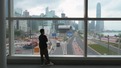 a man looks out of a window to the view of the hong kong island city skyscrapers and financial district skyline seen in the background