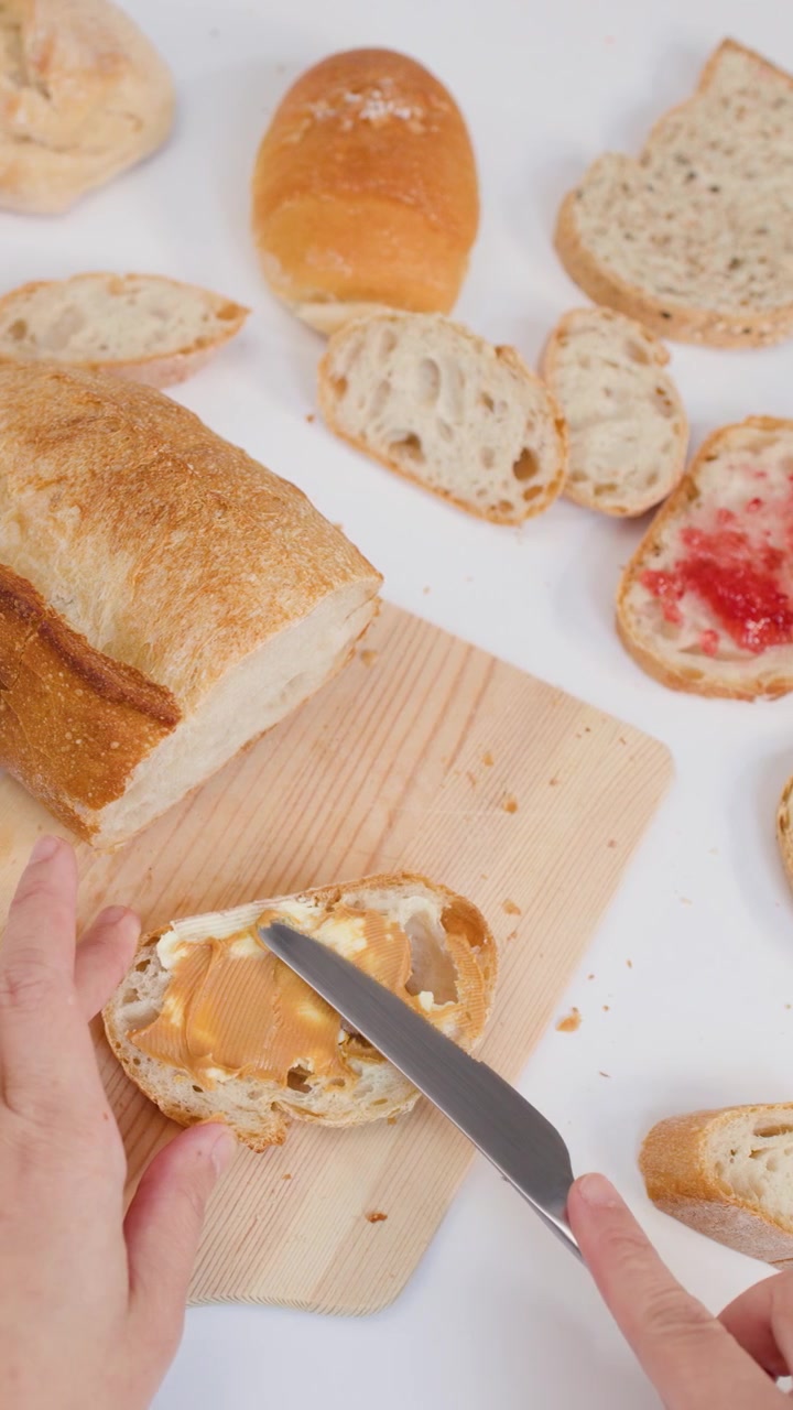 Hands use a knife to spread peanut butter on sourdough bread over a wooden board.