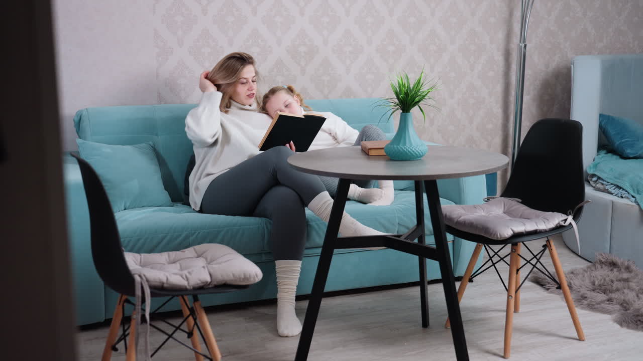 Mother and daughter sitting on blue sofa together reading book in cozy home interior with table, chairs, lamp, cushions, and green plant, sharing quiet bonding moment filled with love