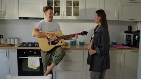 happy middle aged man playing guitar for his pregnant brunette wife in the kitchen in the morning