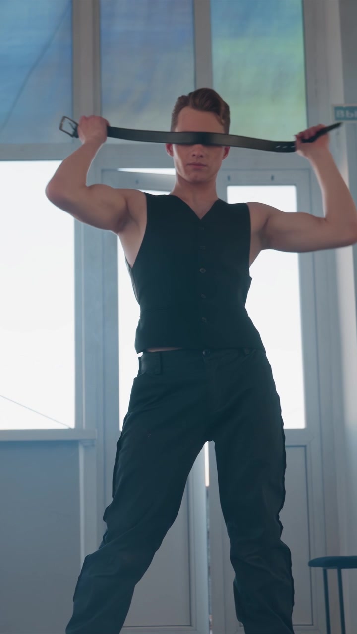 Jovem de colete preto sem mangas ajusta e manuseia alças. O homem manipula objetos perto do corpo, levantando-os. O quarto luminoso apresenta portas brancas e painéis de vidro fosco.