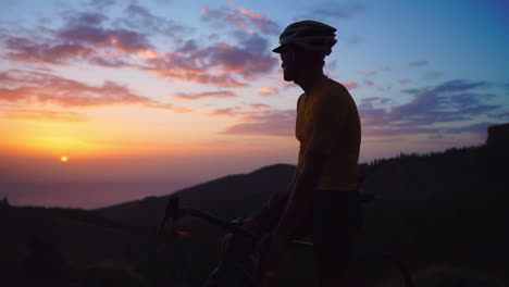 young athlete in yellow t-shirt, helmet, and gear rests on mountain peak, gazing at mountains and setting sun post-training