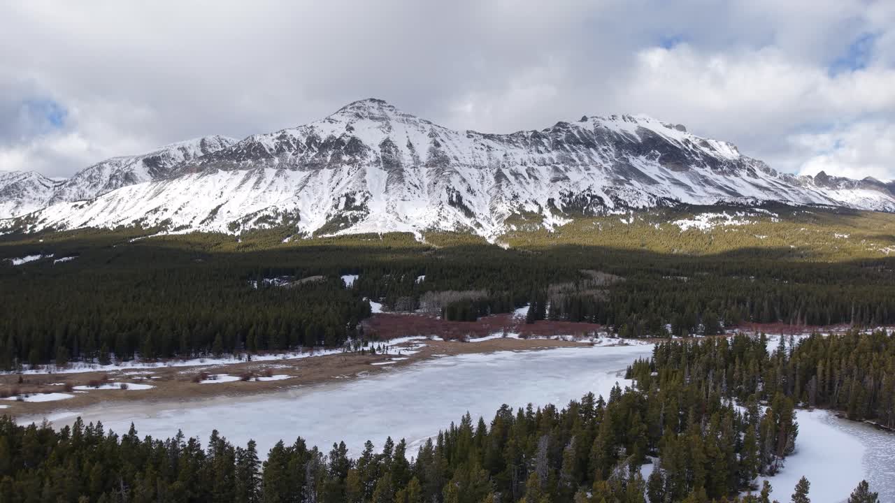 Riprese aeree con drone che mostrano una catena montuosa innevata vicino a Whitefish, Montana, con foreste di conifere e un fiume tortuoso alla sua base sotto un cielo invernale nuvoloso.