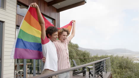 Happy-diverse-gay-male-couple-holding-rainbow-lgbt-flag-on-balcony-at-home,-slow-motion