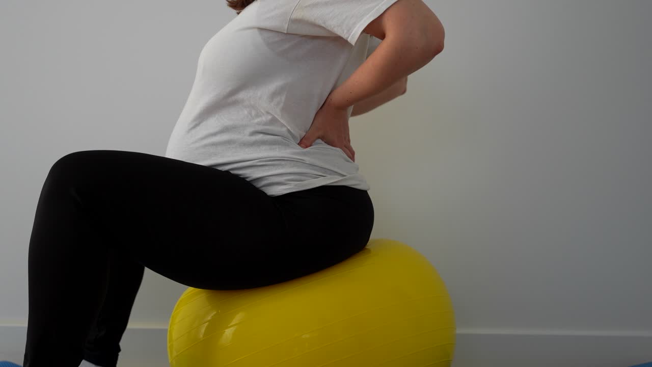 Pregnant woman sits on fitness ball and stretches her back for pain relief