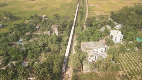 aerial orbit of a train gliding through a rural village in bangladesh, surrounded by greenery and peaceful countryside life.