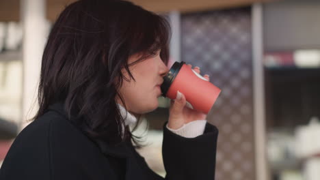 vista lateral de una mujer con abrigo negro bebiendo café de una taza roja al aire libre, expresión relajada con un fondo borroso de entorno urbano con patrones y detalles arquitectónicos