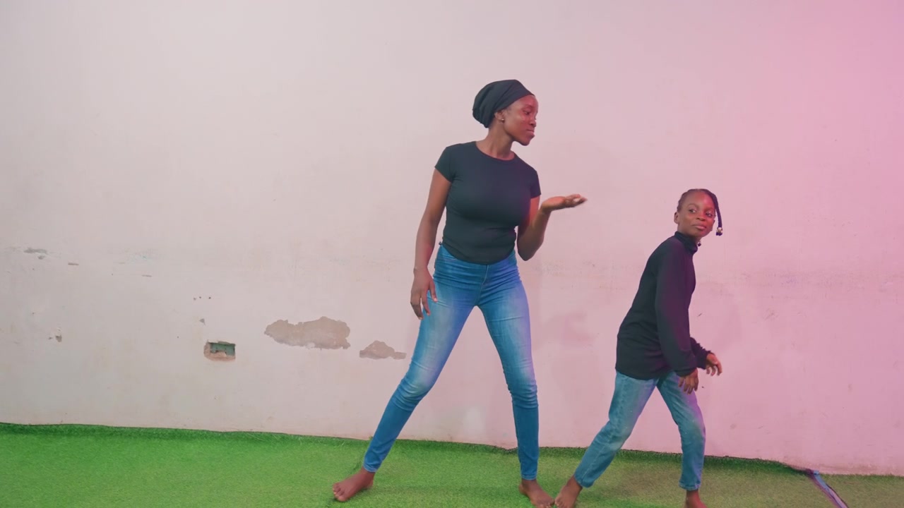 Black mother and child dancing together, energetic duet practice with synchronized steps, full body movement on green turf, pastel wall backdrop, denim fashion, barefoot freedom, rhythmic smiles