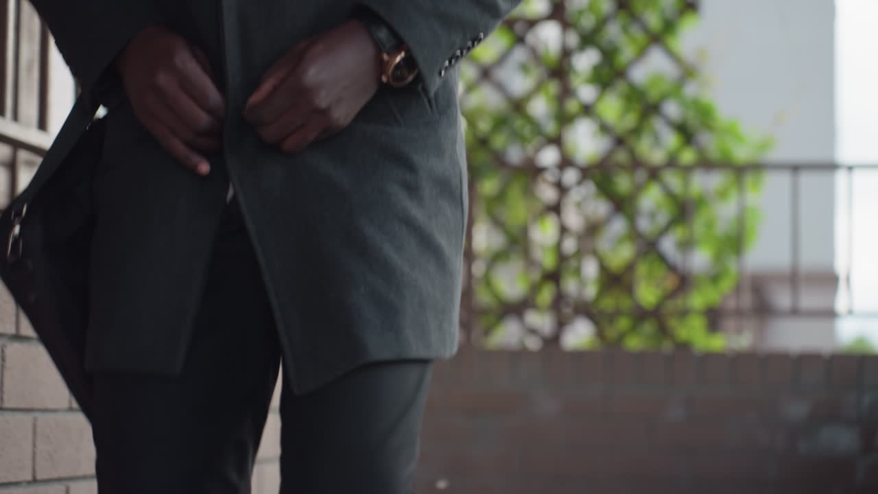 Vista de la cintura que muestra una mano abrochando un abrigo de lana oscura, los dedos cerrando el botón cerca del ojal, un reloj de pulsera brillante, pantalones negros visibles, la correa del bolso colgada y una celosía con hojas frescas.