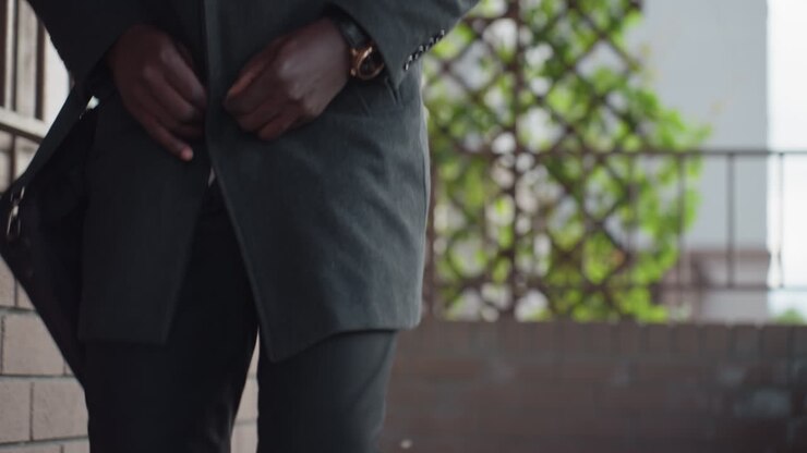 waist view shows hand figure fastening dark wool coat, fingers closing button near buttonhole, wristwatch gleaming, black trousers visible, shoulder bag strap hanging and lattice with fresh leaves