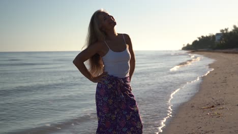 Una-Mujer-Bonita,-Madura-Y-Retroiluminada-En-Una-Playa-Mira-Hacia-Otro-Lado-Del-Mar-Y-Luego-Levanta-Los-Brazos-Y-Sonríe-Mirando-Al-Cielo