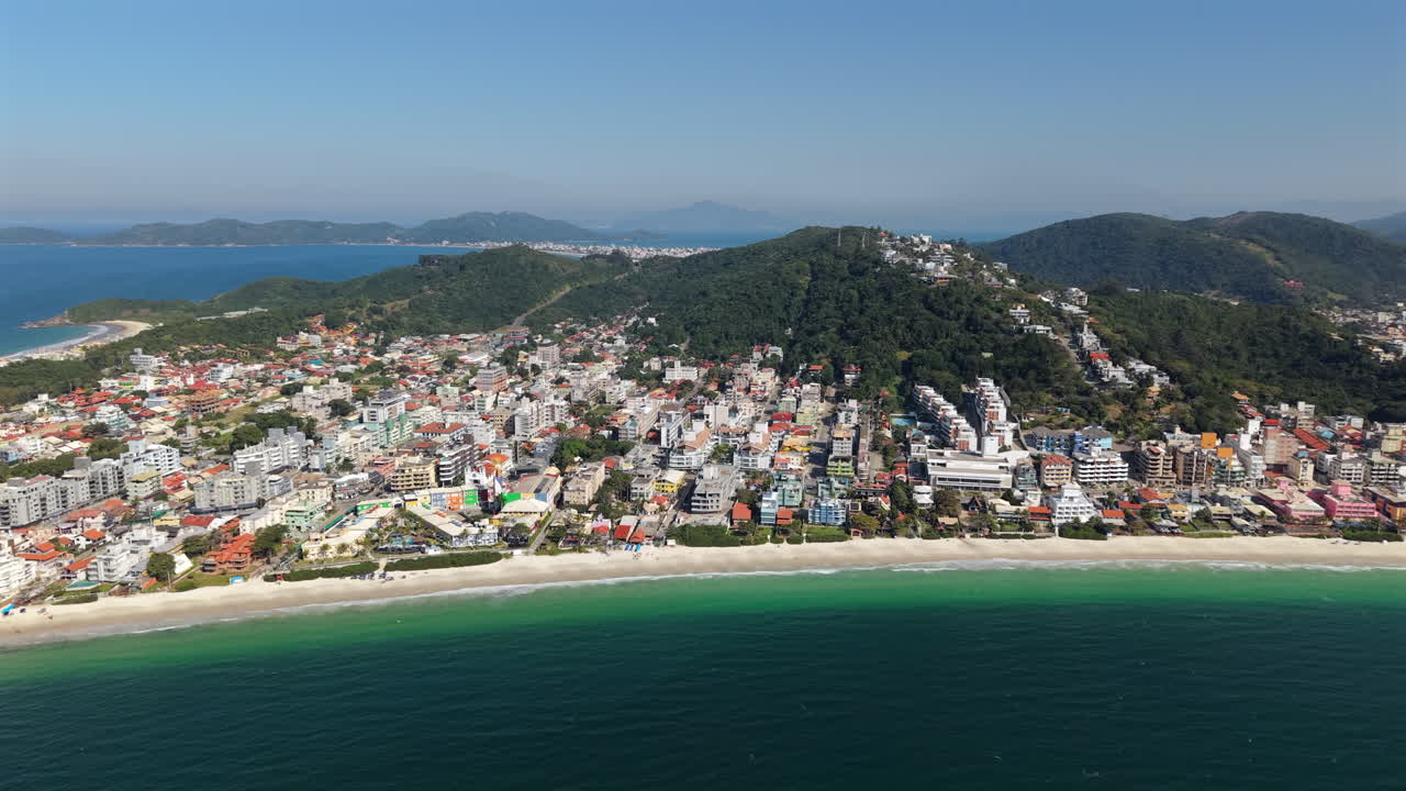 Bombinhas, Santa Catarina, Brazil is shown from above, revealing colorful buildings, a sandy beach, and the vast ocean, creating a vibrant and inviting coastal scene