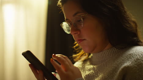 close up of young woman spending evening at home sitting on sofa with mobile phone scrolling through internet or social media