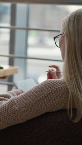 vista trasera de una mujer sentada en una mesa con un libro y una taza de café, escribiendo pensativamente en un cuaderno, mostrando una atmósfera acogedora y enfocada, con un fondo suave de una cafetería o espacio de oficina