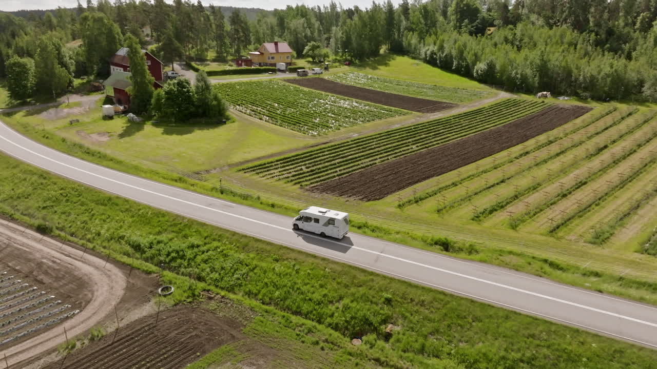 Drone che segue un camper che guida tra fienili e campi rurali, estate