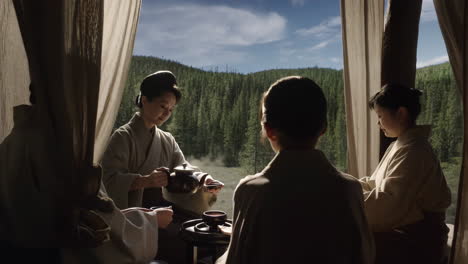 women in traditional attire engaging in a tea ceremony with a forest view