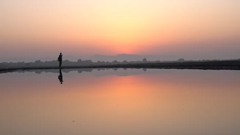 Silhouette-Of-Man-Standing-With-A-Perfect-Reflection-On-The-Salt-Pan-During-Sunset-Time