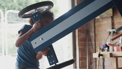 side view of pre teen boy standing at a workbench in the garage and turning over his racing kart, backlit