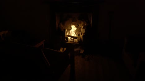 Una-Silla-Vieja-Está-Iluminada-En-Una-Habitación-Oscura-Por-La-Luz-Parpadeante-De-Un-Fuego-De-Leña-En-Una-Chimenea-Vieja