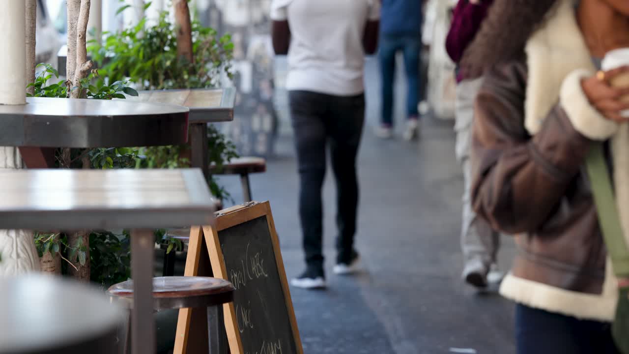 Forbipasserende går forbi en kafé, med et skarpt krittskilt i fokus, mens en kvinne i saueskinn med kaffe går forbi