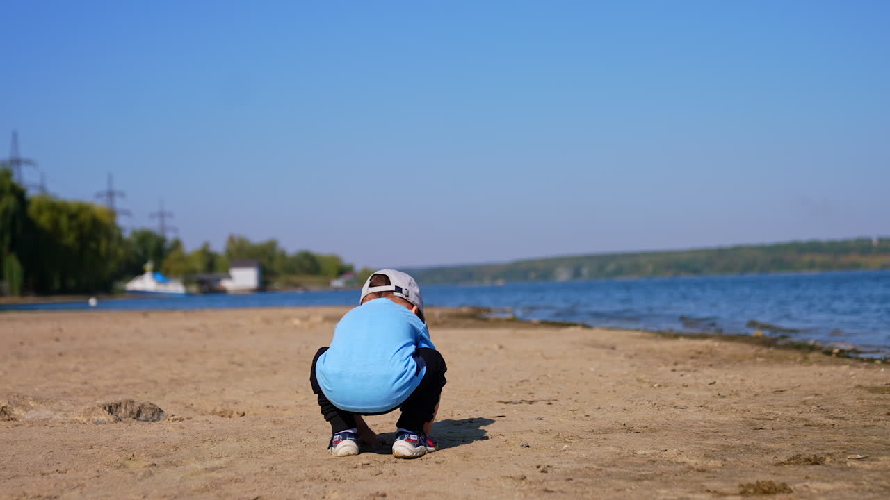 Toddler boy sits squatted on the sunny beach. Rear view of a baby playing in the sand in summer.