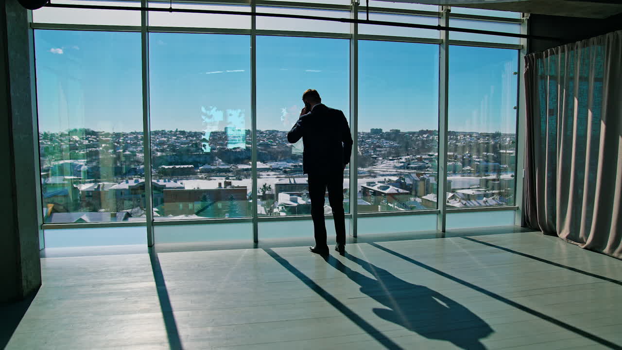 Top manager dealing with business partners through the phone. Male entrepreneur in elegant suit standing by the window and talking the phone. Panoramic windows view.