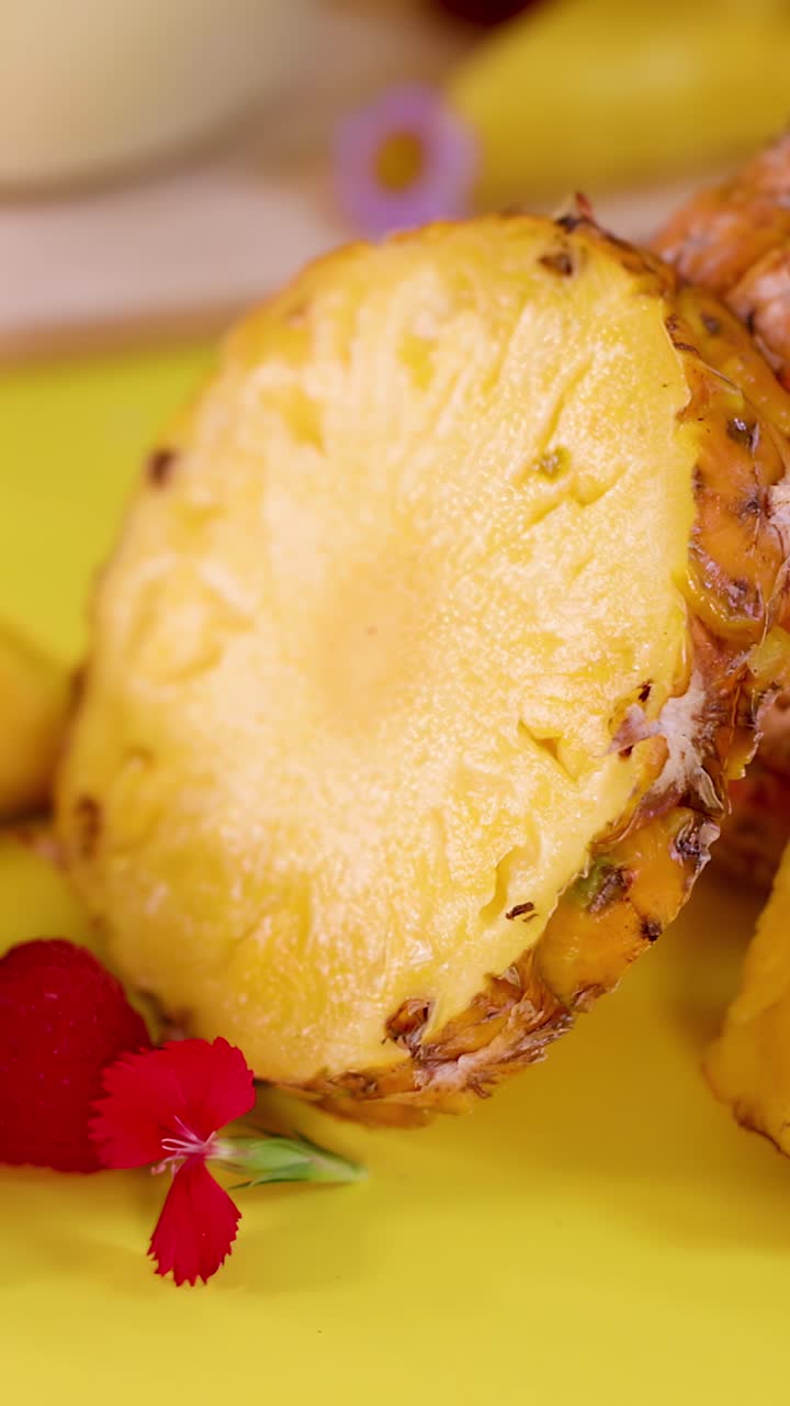 Close-up rotating pineapple slices with strawberries, raspberries, flowers on vibrant yellow studio background