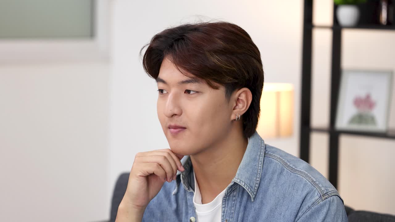 A young Asian man in a denim shirt sits thoughtfully, resting his chin on hand