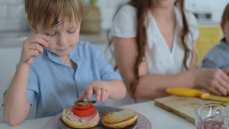 Una-Familia-Feliz-Es-Una-Madre-Joven-Y-Hermosa-Con-Un-Vestido-Blanco-Y-Dos-Hijos-Con-Camisas-Azules-Preparando-Una-Cocina-Blanca-Juntos-Cortando-Verduras-Y-Creando-Berger-Saludable-Para-Los-Niños.