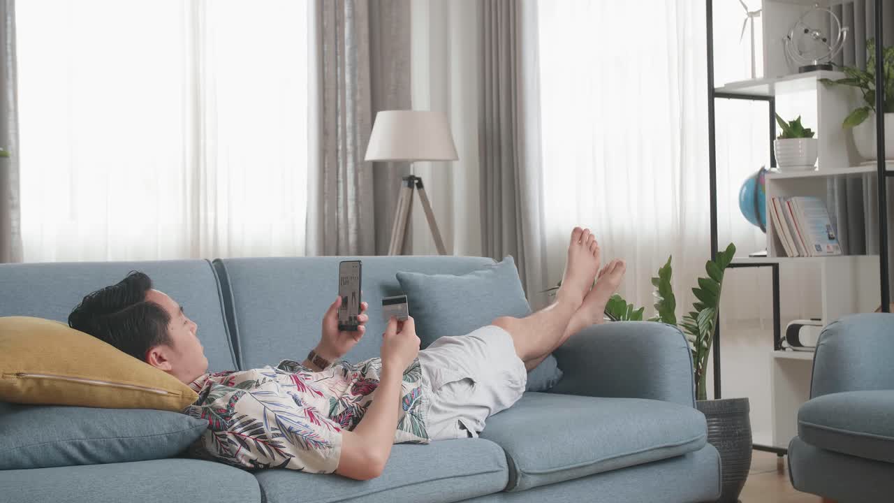 Asian Man Lying On Couch In Living Room Holding Credit Card And Using Smartphone Buy Fashion Clothes Online