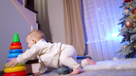 cute smiling baby in white sport suit drops on the floor and crawls up to the colorful toy pyramid. child plays at home on christmas holidays.