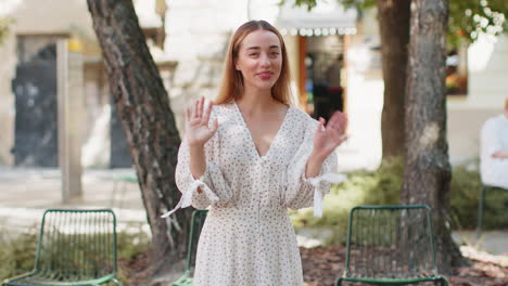 woman in dress smiling friendly at camera waving hands gesturing hi greeting goodbye on city street