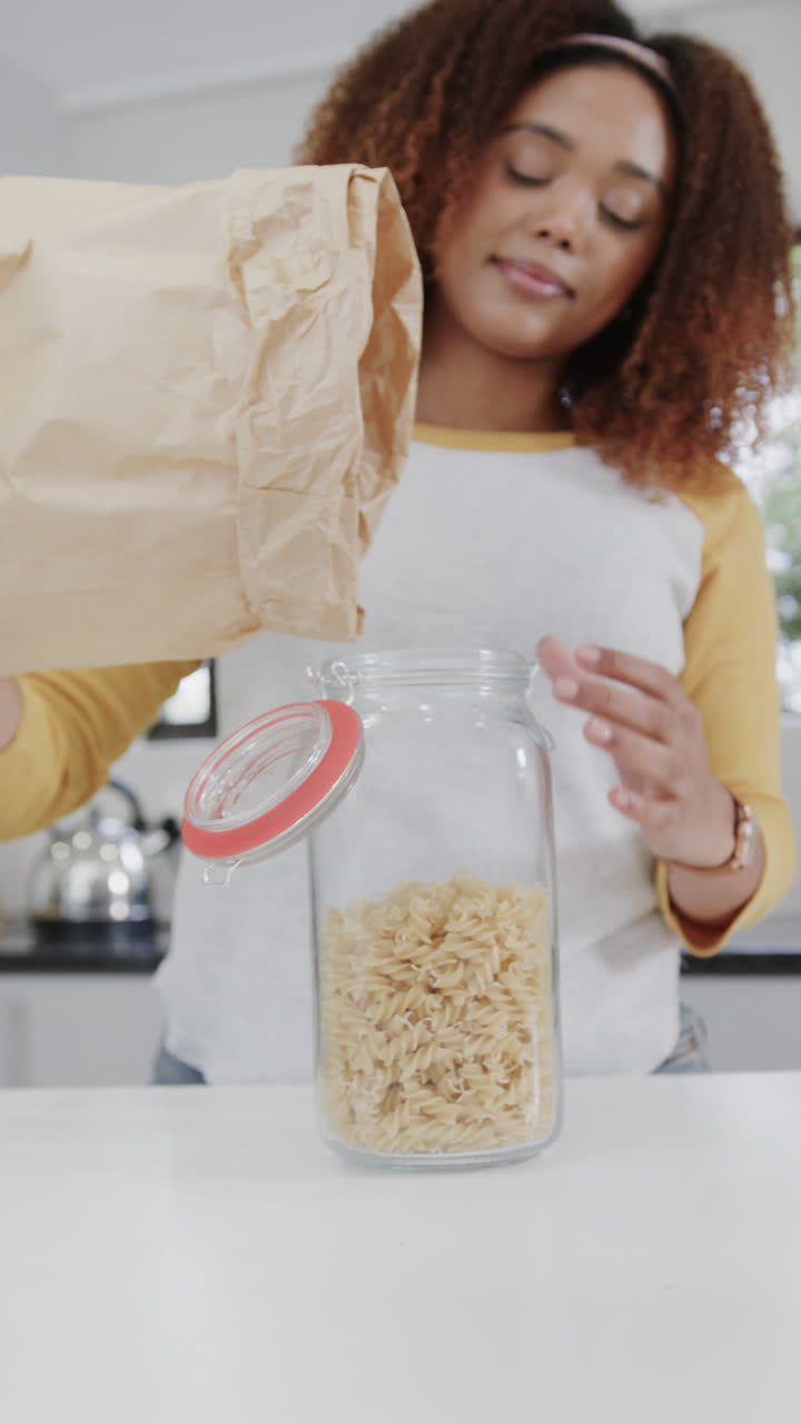 Vertical video of happy african american woman pouring pasta into jar in kitchen in slow motion
