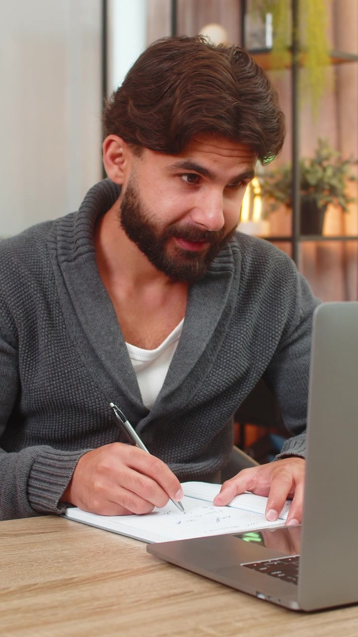 Indisk mann studerer online på bærbar PC og noterer fra e-bok til notatbok med fokusert læring og rolig stemning