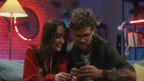 Joven-Feliz-Novio-Y-Novia-Sentados-En-El-Sofá-Por-La-Noche-Y-Mirando-La-Pantalla-Del-Teléfono-Inteligente-Mientras-Hablan