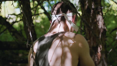 back view of young man in headband punching bark on tree trunk with rhythmic shoulder strikes, muscles tensing against rough wood, surrounded by tangled branches and filtered sunlight in dense forest