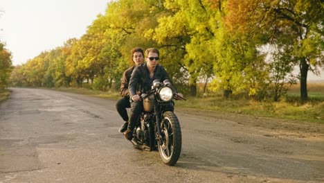 Tracking-shot-of-handsome-man-in-sunglasses-riding-a-motorcycle-with-his-girlfriend-behind,-traveling-together-on-the-asphalt