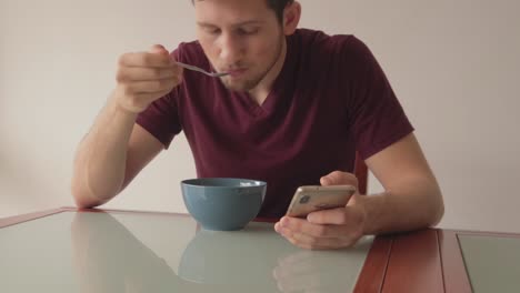 Young-man-sitting-at-table-looking-at-phone-while-eating-cereals,-still-shot-in-minimalistic-setup