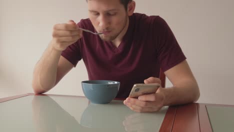 young man sitting at table looking at phone while eating cereals, still shot in minimalistic setup