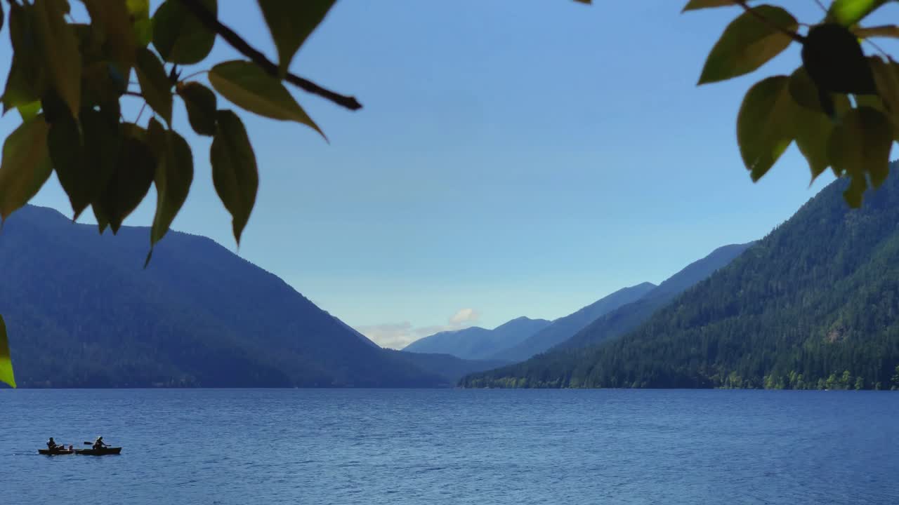 hermosa escena del lago con montañas en el fondo, hojas en primer plano, dos personas flotan en el marco de una canoa