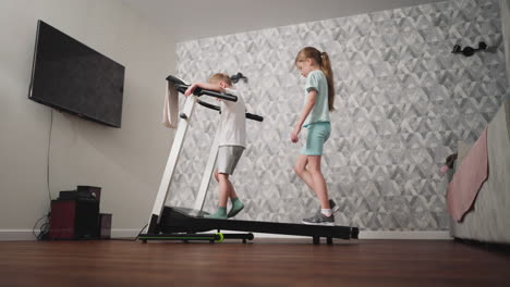 side view of young boy holding handle for balance while walking on treadmill as girl cautiously follows behind, maintaining careful steps during indoor fitness activity in modern living room