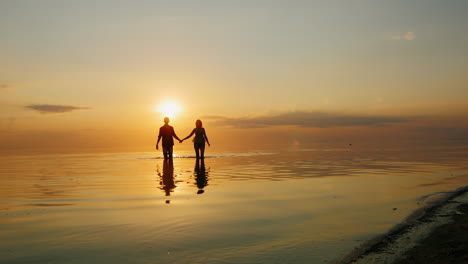 Mann-Und-Frau-Laufen-Bei-Sonnenuntergang-Durch-Das-Wasser-Zum-Kamera-Fliegenspray