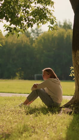 woman sitting thoughtfully by large tree in sunlit park, sunlight casting gentle shadows around her, background includes lush green trees, distant football goalpost, and car in motion