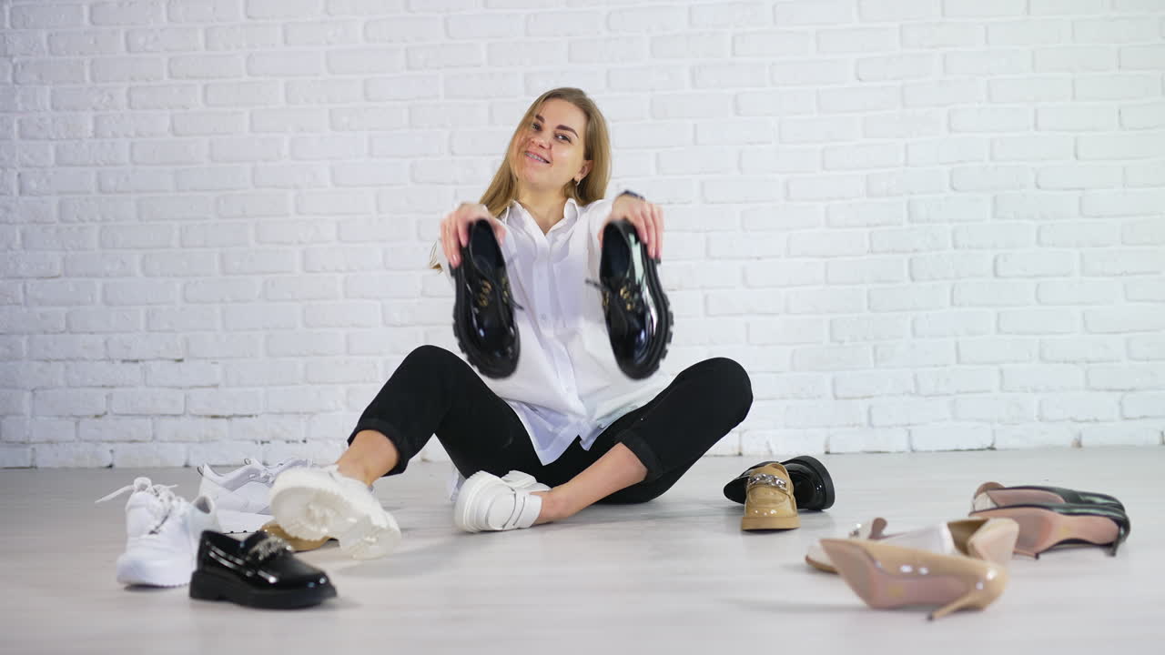 Young long-haired lady demonstrating new shiny black shoes with laces. Woman among the footwear at white background.