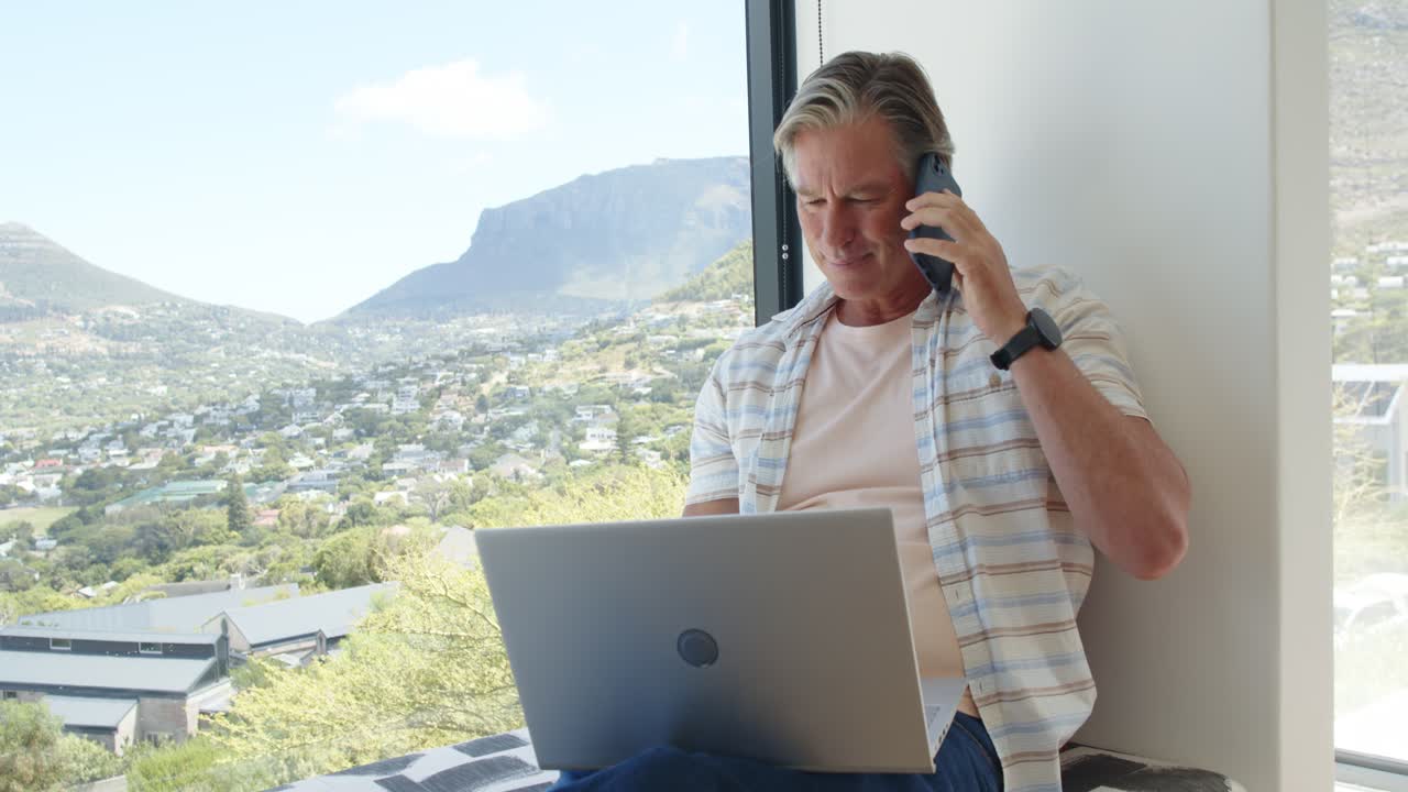 Mature man placing laptop on lap, holding phone to ear and typing for work, copy space