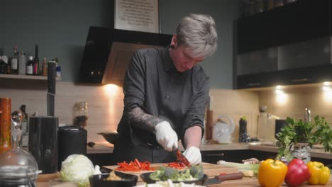 Young-professional-male-chef-in-an-elegant-black-shirt-with-an-alternative-look,-tattoos,-gloves-cutting-red-peppers-A-modern,-fancy-looking-kitchen-with-fresh-vegetables-besides-Cinematic-Sliding-4K