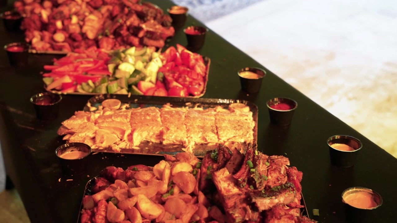 Elegante tavolo da buffet presenta un assortimento di carni alla griglia, verdure arrosto, insalate fresche e salse disposti su vassoi neri sotto un'illuminazione calda. Perfetto per riprese di cene gourmet