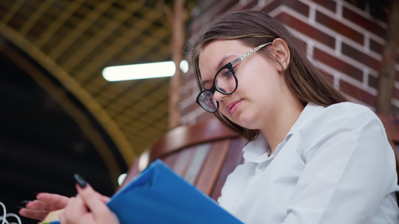 joven con gafas lee un libro mientras está sentada en un centro comercial moderno. pilar de ladrillo detrás de ella y luces bokeh en el fondo añaden profundidad, está enfocada, comprometida e inmersa en su lectura