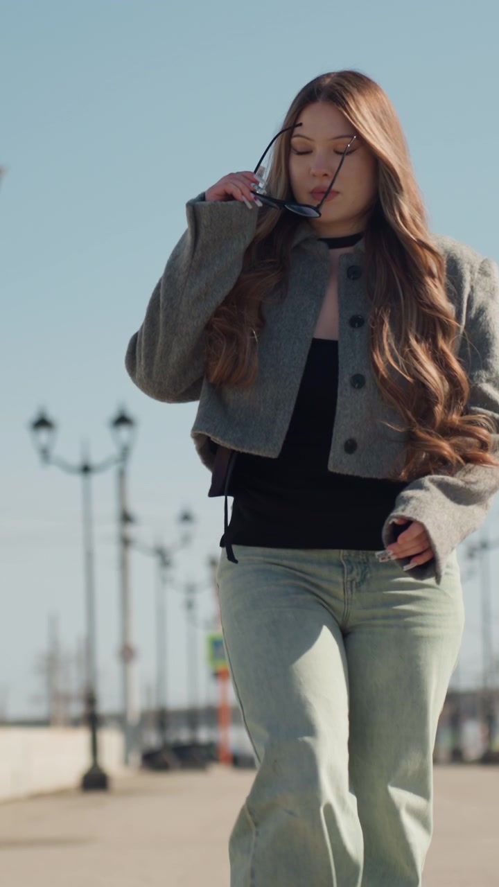 Krásná žena kráčející po ulici směrem k fotoaparátu, s sebevědomým modelem s vlajícími vlasy, džínovými kalhotami a zkrácenou bundou, gestem se slunečními brýlemi, slunný bulvár a módní postoj.